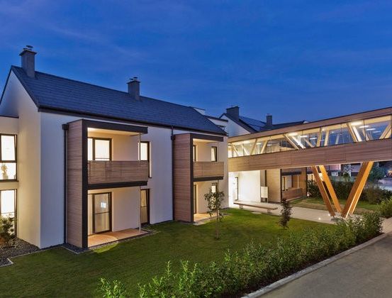 Ein modernes Mehrfamilienhaus mit einer Glasbrücke, die zwei Gebäude bei Einbruch der Dunkelheit verbindet.
