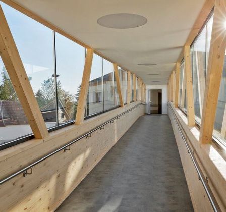 Ein langer Korridor mit Holzwänden und Glasfenstern. Sonnenlicht scheint durch die Fenster. Der Korridor ist leer mit einem Betonboden.