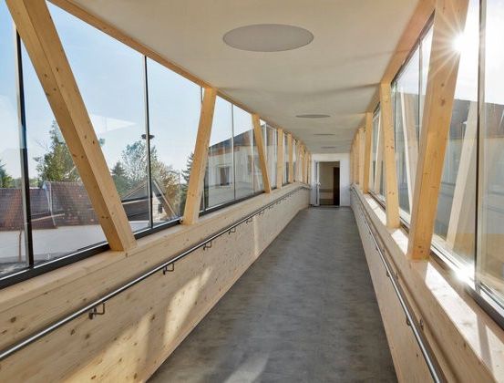 Ein langer Korridor mit Holzwänden und Glasfenstern. Sonnenlicht scheint durch die Fenster. Der Korridor ist leer mit einem Betonboden.