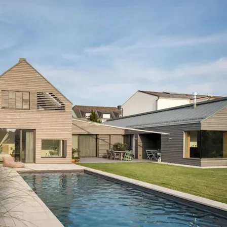 Ein modernes Haus mit großem Swimmingpool, hölzerner Außenfassade und einer Terrasse mit Stühlen und Tischen. Das Haus hat große Fenster und ist von grünem Rasen umgeben.