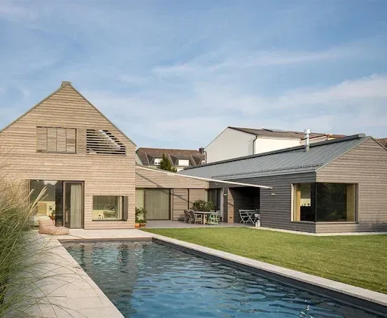 Ein modernes Haus mit großem Swimmingpool, hölzerner Außenfassade und einer Terrasse mit Stühlen und Tischen. Das Haus hat große Fenster und ist von grünem Rasen umgeben.