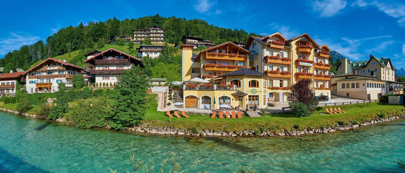 Ein großes gelbes Hotel namens Hallenbad befindet sich am Hang mit einem Fluss davor, umgeben von Bäumen und anderen Gebäuden.