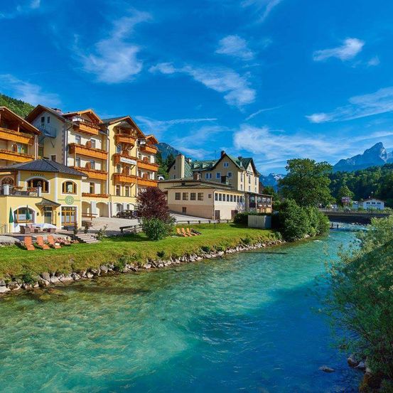 Ein ruhiges Hotelkomplex mit Balkonen und Liegestühlen an einem klaren Fluss unter einem blauen Himmel mit Bergen im Hintergrund.