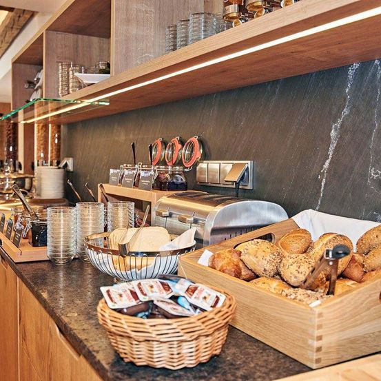 Ein Frühstücksbuffet mit Brot, Gewürzen und Besteck. Ein Toaster und ein Regal mit Gläsern und Gläsern. Eine schwarze Wand mit einem Lichtstreifen.