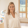 Eine Frau steht lächelnd in einem Raum mit Holzboden. Sie trägt eine weiße Bluse und Jeans und steht vor einem Schreibtisch mit einer Globus und Spielzeug. Ein großes Fenster mit Holzrahmen befindet sich hinter ihr.