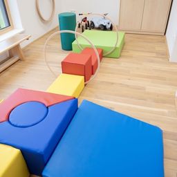 Ein Spielzimmer mit Holzboden, bunten Kissen und einer Hula-Hoop. Ein Fenster, ein Holzbank und ein Spielzeugauto sind ebenfalls vorhanden.