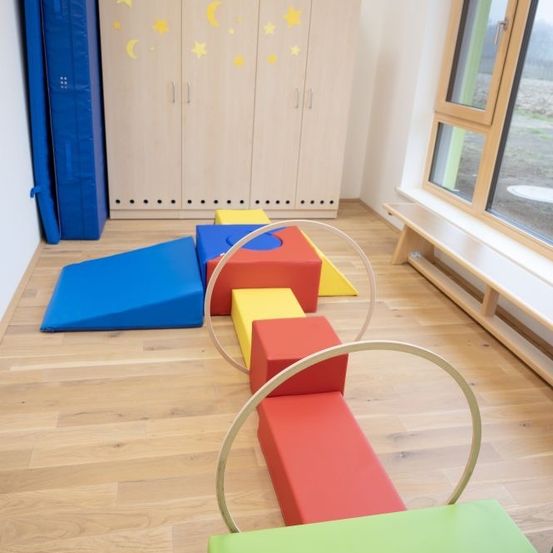Ein Spielzimmer mit Holzboden, bunten Bauklöcken, Matten und einer Holzbank am Fenster.