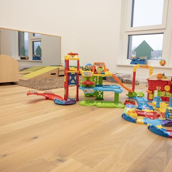 Ein Spielzimmer mit Holzboden und verschiedenen farbigen Spielzeugen, darunter ein großes Spielzeug-Zugset und ein kleines Spielhaus.