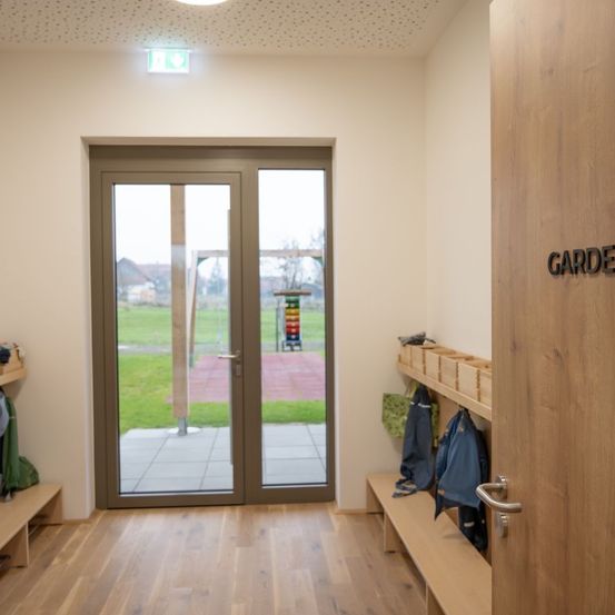 Ein Raum mit Holzboden, einer Glastür zu einer Rasenfläche und einer Holzbank, an der Kleidung hängt.