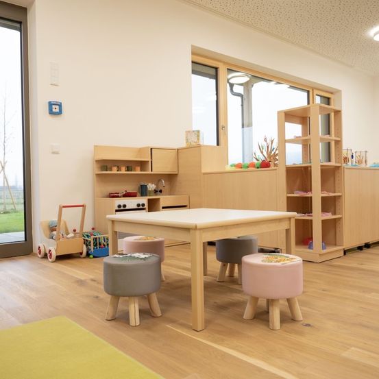 Ein Kinderspielzimmer mit Holzboden und Möbeln, bestehend aus einem Tisch, Hockern, Regalen und Spielzeug. Es gibt eine Glastür, die zu einem Rasenbereich führt.
