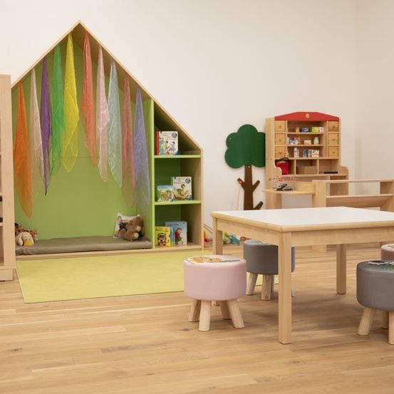 Ein Spielzimmer mit einem bunten Haus, einem Tisch und Hockern. Das Haus hat bunte Schals, die von der Dachspitze hängen, und einen Teppichboden. Ein kleiner Bücherregal und ein Baum sind ebenfalls im Zimmer.