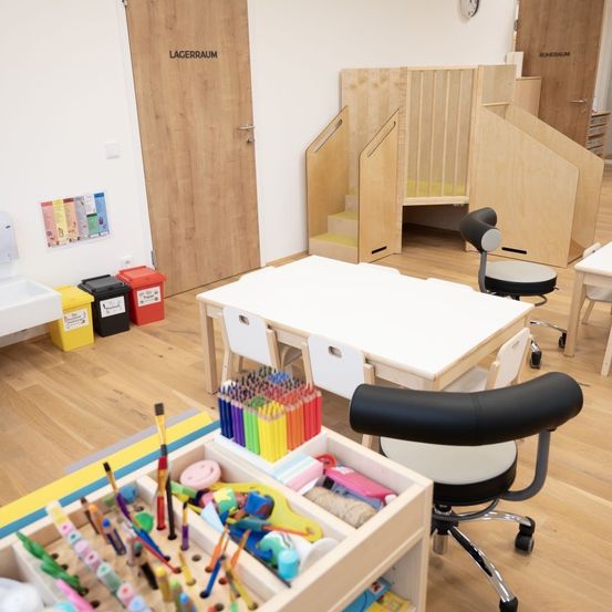Ein Kinderspielzimmer mit Holzböden und einem weißen Tisch voller bunter Stifte. Der Raum hat eine Tür mit der Aufschrift 'Lagerraum' und eine Holzstruktur mit Treppen. Abfallbehälter stehen an der Wand.