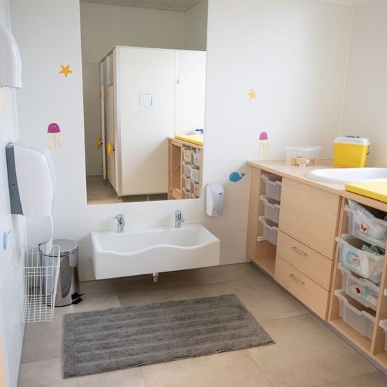 Ein sauberes Badezimmer mit einem weißen Waschbecken, einem Spiegel und hölzernen Schränken. Ein Mülleimer, eine Matte und Aufbewahrungsboxen sind in der Nähe.