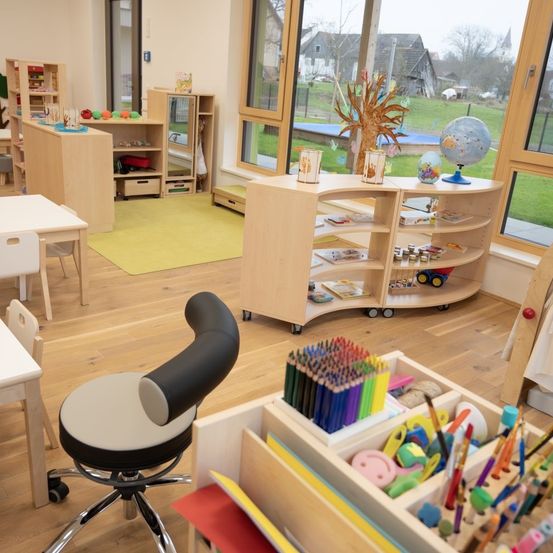 Ein Raum mit Holzboden, ausgestattet mit einem Globus, einem Regal mit Büchern und Spielzeug, einem Stuhl, einem Tisch und großen Fenstern mit Blick aufs Freie.