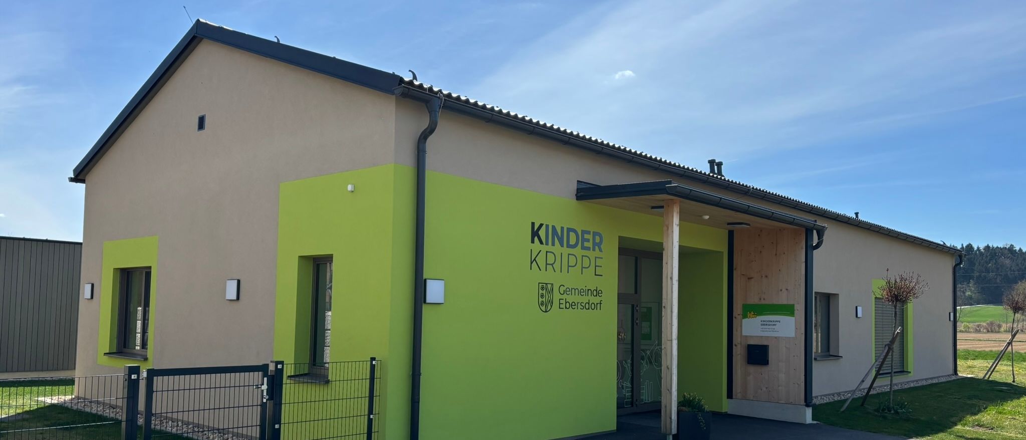 Die Vorderansicht eines Gebäudes mit grünen Wänden, einem schwarzen Dach und einem grünen Rasen. Das Gebäude hat die Worte 'KINDER KRIPPE' und 'Gemeinde Ebersdorf' an der Wand geschrieben.
