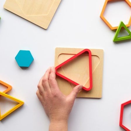 Eine Hand hält eine rote dreieckige Aussparung auf einer Holzplatte mit verschiedenen farbigen geometrischen Formen darum herum.