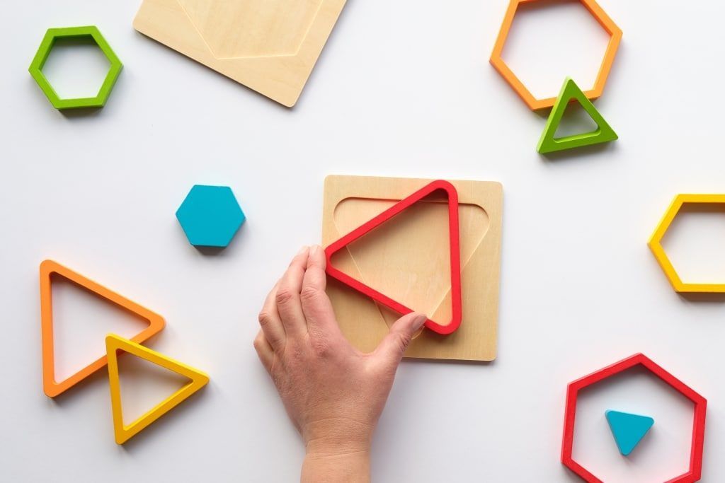 Eine Hand hält eine rote dreieckige Aussparung auf einer Holzplatte mit verschiedenen farbigen geometrischen Formen darum herum.