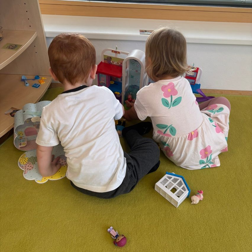 Zwei Kinder sitzen auf dem Boden und spielen mit Spielzeug. Ein Junge schaut sich ein Puppenhaus an, und ein Mädchen spielt mit einer Puppe.