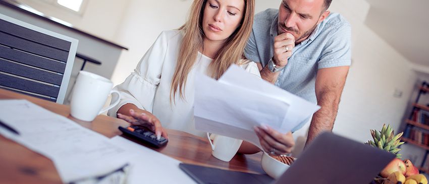 Ein Paar schaut sich Rechnungen an und benutzt einen Taschenrechner, während es in einer Küche an einem Schreibtisch sitzt.