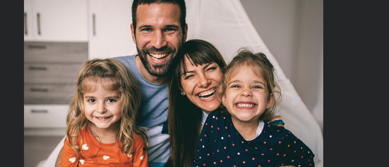 Eine vierköpfige Familie lächelt für ein Foto, mit zwei Mädchen und einem lächelnden Vater und Mutter. Die Mutter trägt Ohrringe und die Mädchen starr-gemusterte Kleidung.