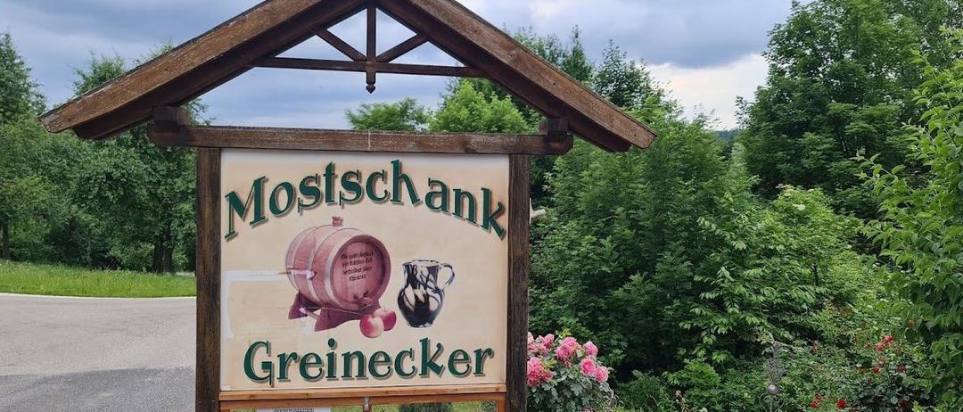 Ein hölzernes Schild mit einem dreieckigen Dach steht in einem Garten. Es steht 'Mostschank Greinerecker' und zeigt einen Fass und Krug. Unten steht ein kleineres Schild mit 'MONTAG BIS DONNERSTAG MITTWOCH FREITAG' und Öffnungszeiten.
