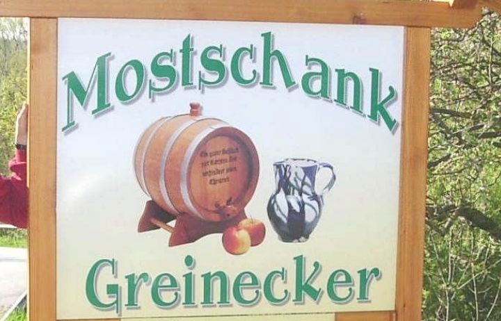 Ein Schild für Mostschank Greinerecker zeigt einen Fass, eine Kanne und Äpfel. Es enthält Öffnungszeiten und eine Nachricht, dass es an Feiertagen geschlossen ist.