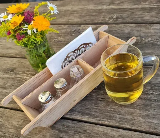 Ein Holztablett mit Salz- und Pfefferstreuer, einer Vase mit Blumen und einer Tasse Tee steht auf einem Holztisch.