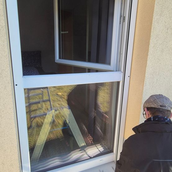 Ein Mann reinigt ein weißes Fenster mit weißem Rahmen. Das Fenster ist offen und ein Reflektion einer Leiter ist sichtbar. Der Mann trägt eine Kappe und eine Brille.