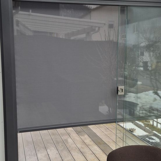 Ein grauer Sichtschutzrollo ist über einer Glastür auf einer Holzterrasse gezogen. Schnee ist auf dem Boden zu sehen.
