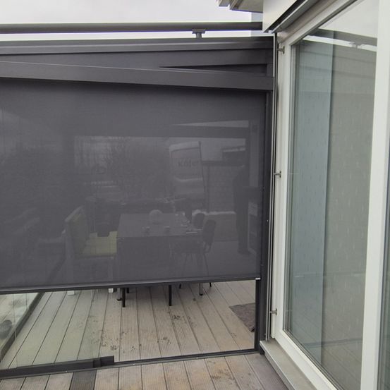 Ein moderner Balkon mit einem Rollo und einem Esstisch mit Stühlen. Der Bereich ist von Glaswänden umgeben.