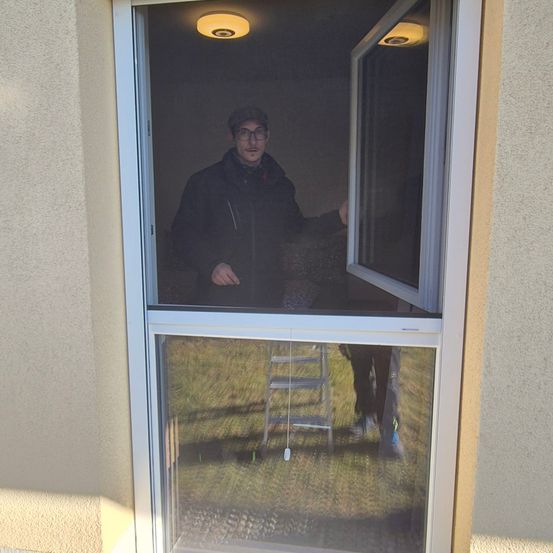 Ein Mann mit Hut und Brille steht in einem Haus und schaut aus einem Fenster. Das Fenster hat einen weißen Rahmen und einen weißen Bildschirm. Draußen steht eine weitere Person in der Nähe einer Leiter.