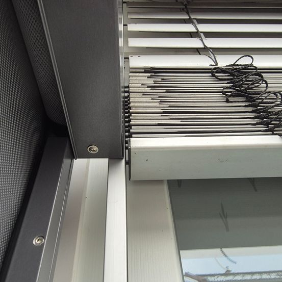 Nahaufnahme eines Fensterrahmens mit einer aufgerollten weißen Jalousie, zeigt Metallhalterungen und eine graue Wand im Hintergrund.