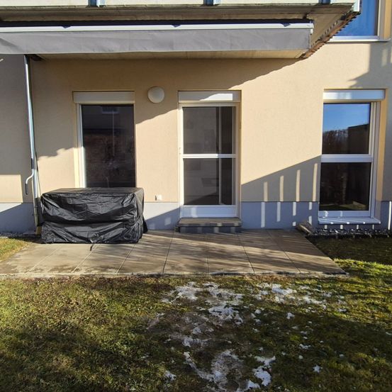 Eine Terrasse mit einem abgedeckten Grill, neben einem beigen Haus mit einer Glastür, einem Fenster und einem überdachten Vordach. Schnee liegt auf dem Boden.