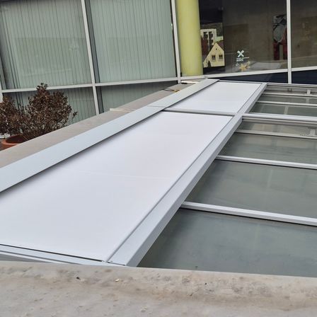 Ein neuer weißer Vordach wird über einem Glasdach auf einem Gebäude mit grünen Jalousien installiert. Links ist ein Topf mit einer Pflanze zu sehen.