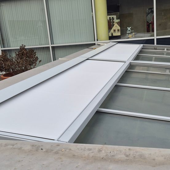 Ein neuer weißer Vordach wird über einem Glasdach auf einem Gebäude mit grünen Jalousien installiert. Links ist ein Topf mit einer Pflanze zu sehen.