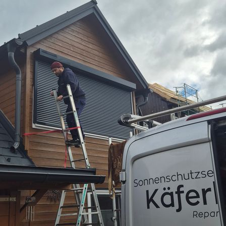 Ein Mann steht auf einer Leiter neben einem Haus mit einer aufgerollten Jalousie. Das Haus hat Holzaußenwände und einen bewölkten Himmel. Ein Van mit der Aufschrift Sonnenschutzse Kaferl steht davor.