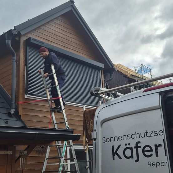 Ein Mann steht auf einer Leiter neben einem Haus mit einer aufgerollten Jalousie. Das Haus hat Holzaußenwände und einen bewölkten Himmel. Ein Van mit der Aufschrift Sonnenschutzse Kaferl steht davor.