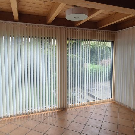 Ein leerer Raum mit Holzbalkendecke, gefliestem Boden und zwei großen Fenstern, die von weißen vertikalen Jalousien bedeckt sind. Draußen befindet sich ein Garten mit Pflanzen und Bäumen.