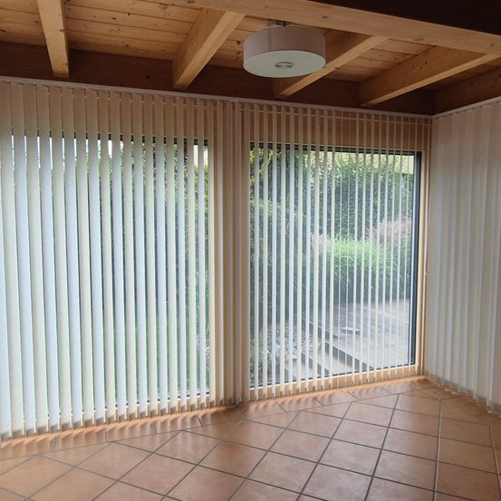 Ein leerer Raum mit Holzbalkendecke, gefliestem Boden und zwei großen Fenstern, die von weißen vertikalen Jalousien bedeckt sind. Draußen befindet sich ein Garten mit Pflanzen und Bäumen.