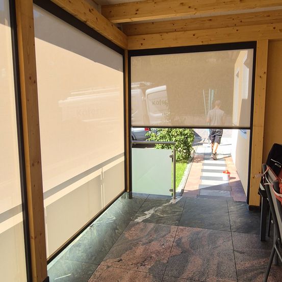 Eine Terrasse mit Holzgeländer, einer Glastür und einer Person, die im Hintergrund geht. Die Terrasse hat einen gefliesten Boden, weiße Vorhänge und ein geparktes Fahrzeug.