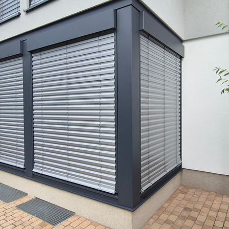 Ein grau verkleidetes Gebäude mit zwei geschlossenen grauen Rollläden. Die Rollläden haben silberne Lamellen. Der Boden ist mit Ziegelsteinen gepflastert.