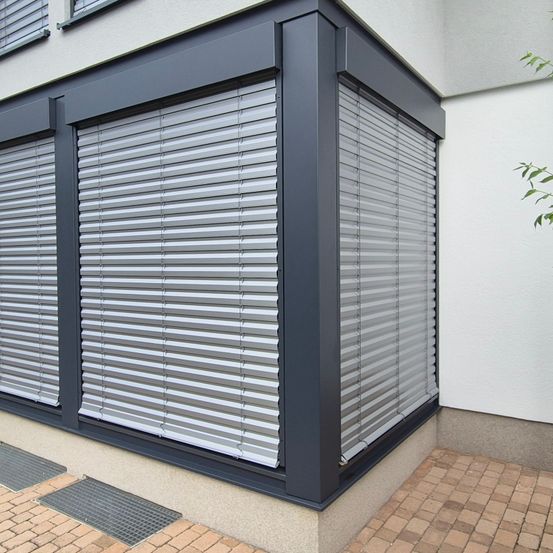 Ein grau verkleidetes Gebäude mit zwei geschlossenen grauen Rollläden. Die Rollläden haben silberne Lamellen. Der Boden ist mit Ziegelsteinen gepflastert.