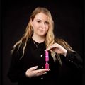 Eine Frau mit blondem Haar lächelt und hält ein rosa zylindrisches Objekt in den Händen. Sie trägt ein schwarzes Langarmshirt und eine Halskette.