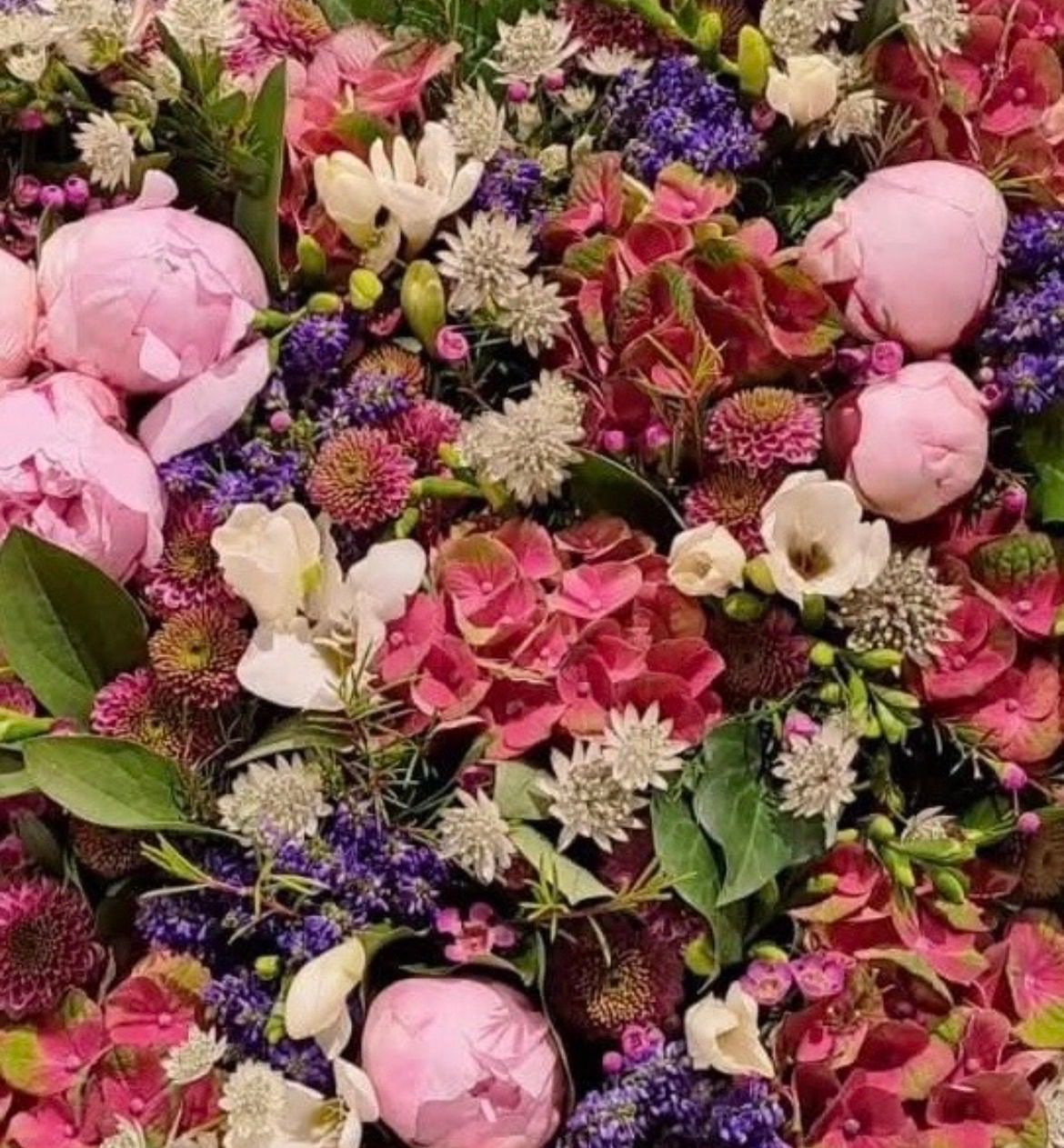 A vibrant arrangement of pink, white, and purple flowers, including peonies and hydrangeas, surrounded by lush green leaves.