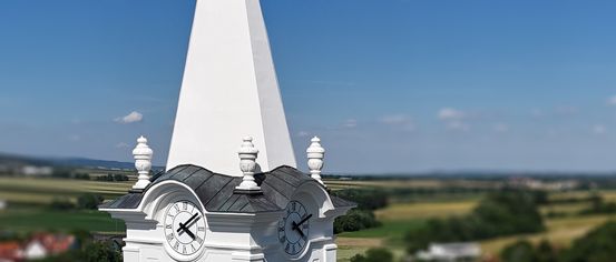 Aerial view of a white church tower with a clock, surrounded by rural houses under a blue sky with clouds.