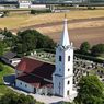 An aerial view of a white church with a red roof and a tall steeple. The church is surrounded by trees and a cemetery.