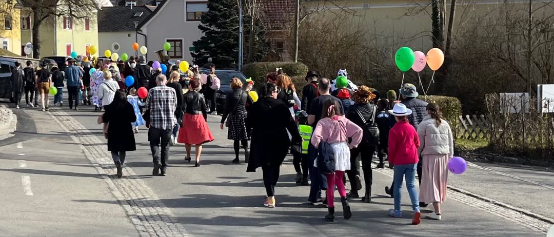 Eine Menschenmenge, einige in Kostümen, geht auf einer Straße entlang, gesäumt von Häusern und Bäumen. Bunte Luftballons zieren die Szene, und der Himmel ist blau mit Wolken.