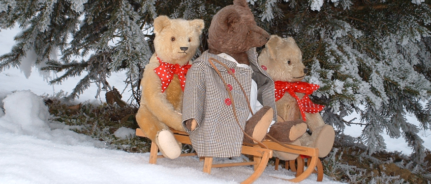 Drei gepolsterte Teddybären sitzen auf einem Schlitten unter einem schneebedeckten Baum, alle tragen rote Schleifen und Winterkleidung.