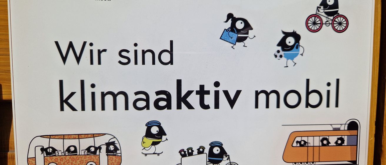 Ein weißes Schild mit schwarzem Text 'Wir sind klimaaktiv mobil' und mehreren Illustrationen von Menschen, die verschiedene Fahrzeuge fahren.