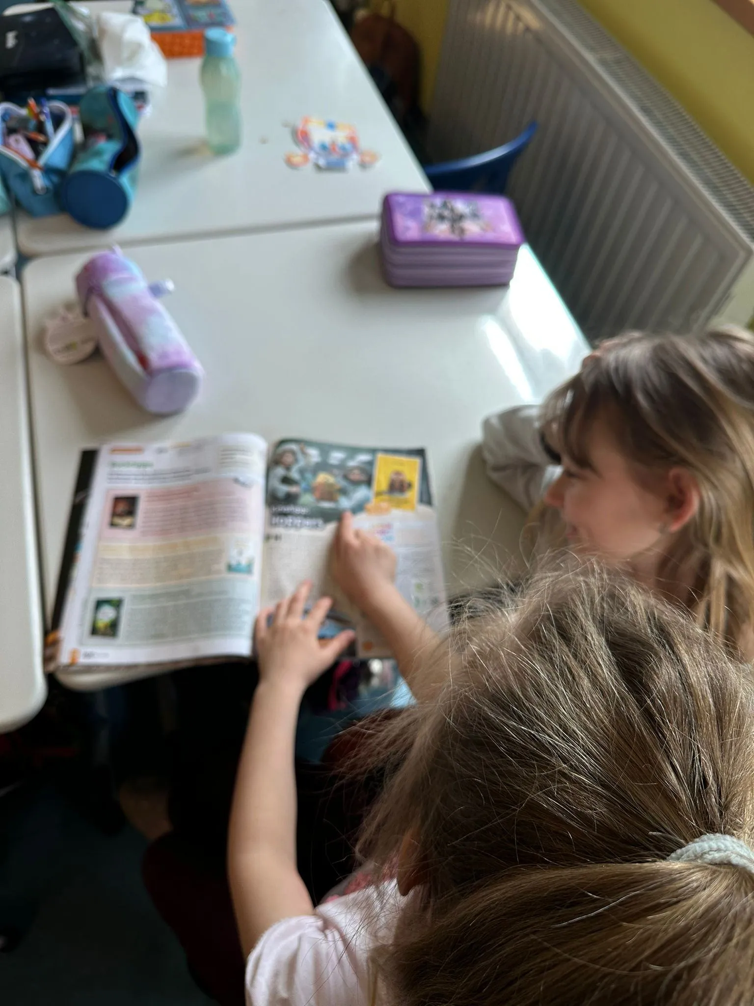 Zwei Mädchen sitzen an einem Schreibtisch mit einem offenen Buch. Eine liest, während die andere auf die Seiten zeigt. Verschiedene Schulutensilien liegen auf dem Schreibtisch.